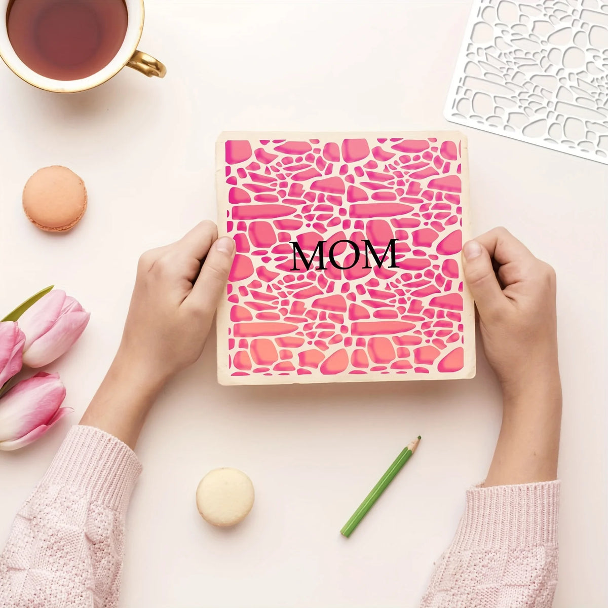 Hands hold a pink stencil card with the word MOM on it, set against a light background.