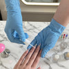 A hand wearing blue nitrile disposable gloves is holding a white tube over another hand on a marble surface.