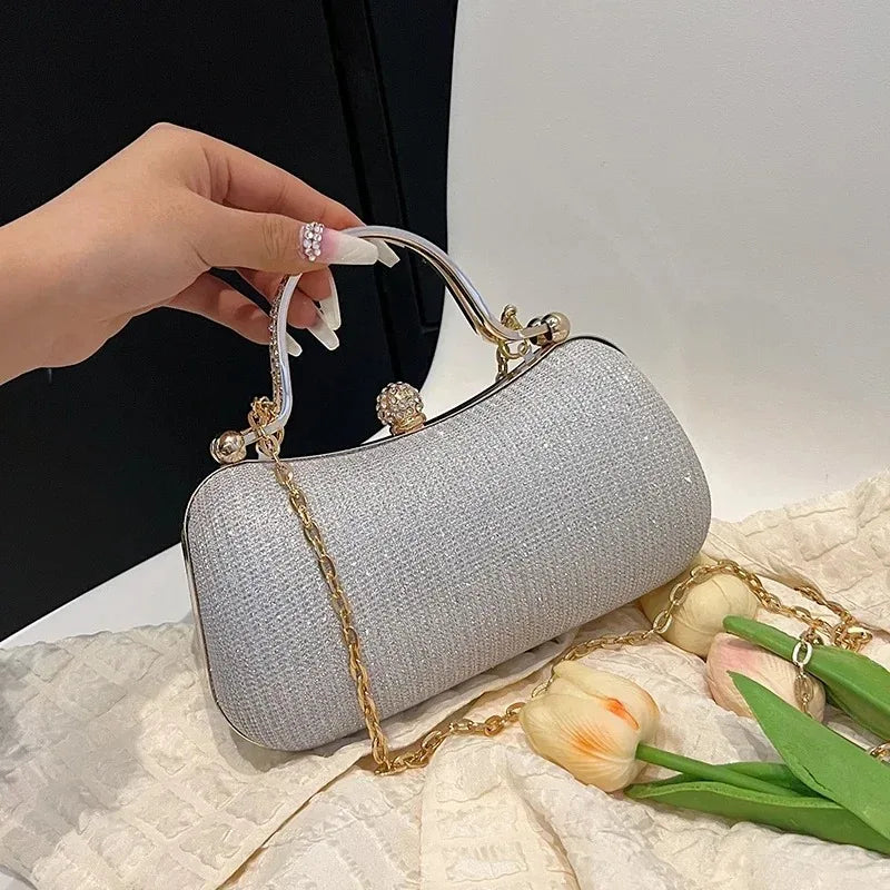 A silver shimmering envelope clutch with a gold chain strap is held by a hand on a light fabric surface.