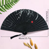 Black folding hand fan with white floral design and a durable handle displayed on a pink background.