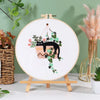 A circular embroidery hoop featuring a black cat and floral designs on white fabric, displayed on a wooden stand.