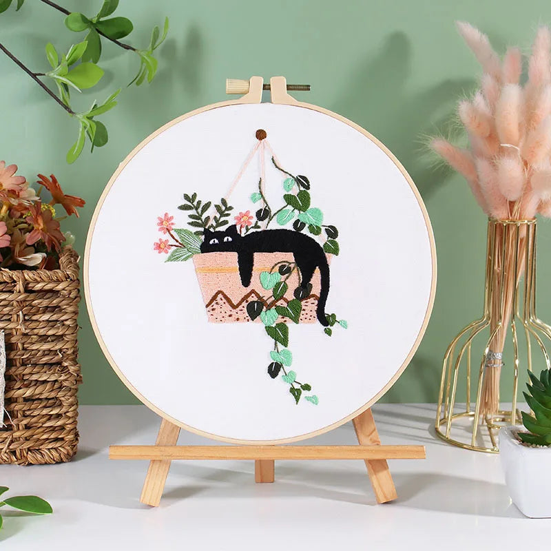 A circular embroidery hoop featuring a black cat and floral designs on white fabric, displayed on a wooden stand.