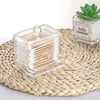 Clear storage dispenser for cotton swabs and pads with a wooden lid placed on a woven mat.