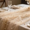 A sheer beige pearl beaded tulle table runner draped elegantly on a white tablecloth for an event.