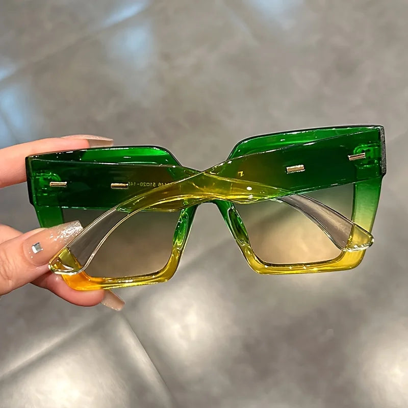A pair of gradient square sunglasses with green and yellow lenses being held by a hand.