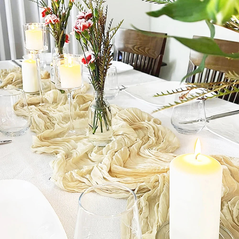 A rustic boho table setting featuring a semi-sheer gauze table runner, candles in glass holders, and greenery.