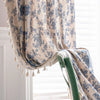 A close-up of a beige cotton-linen retro floral curtain with blue floral patterns and tasseled edges hanging in a window.