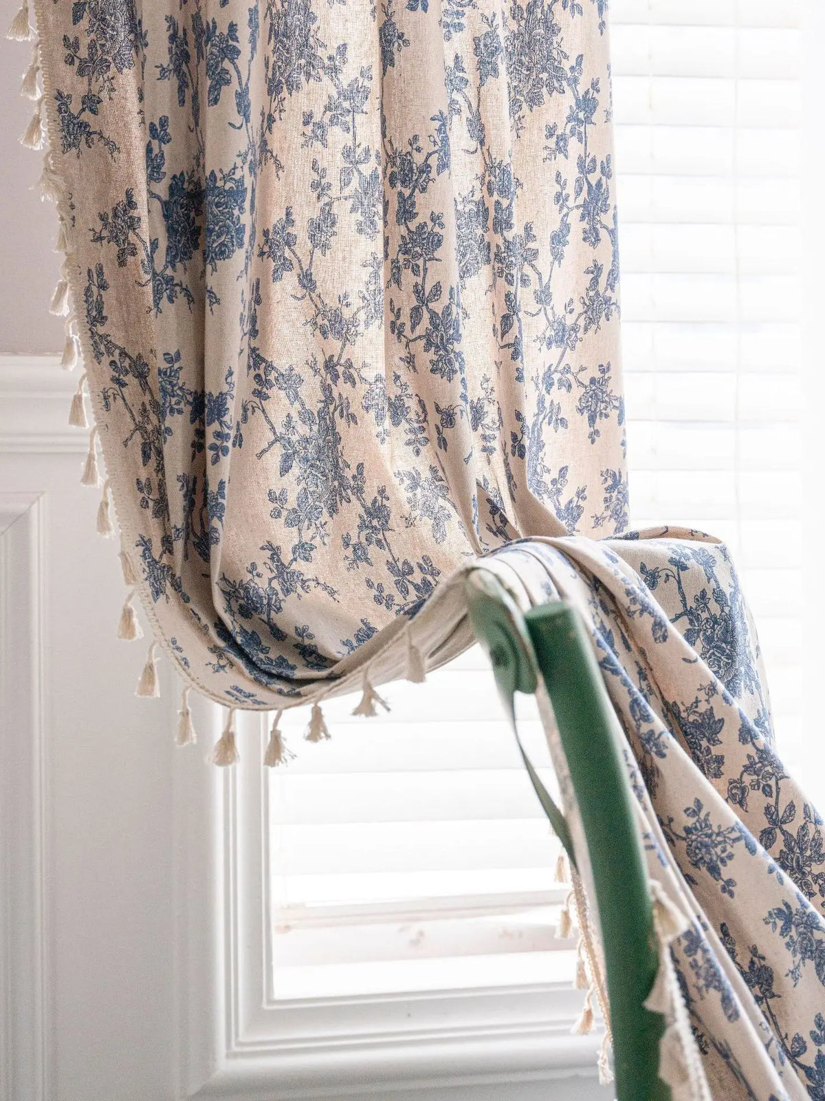 A close-up of a beige cotton-linen retro floral curtain with blue floral patterns and tasseled edges hanging in a window.