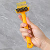 A hand holds an orange dual-sided hairbrush cleaner with rake teeth against a gray background.