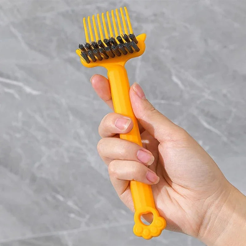 A hand holds an orange dual-sided hairbrush cleaner with rake teeth against a gray background.