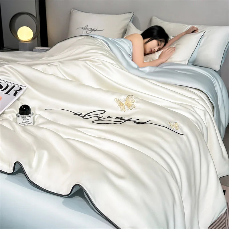 A cream colored ultra-soft comforter with embroidery featuring the words always and a butterfly design, draped on a bed.
