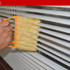 A hand is using an orange tool to clean dust from white horizontal blinds with a yellow dusting brush.