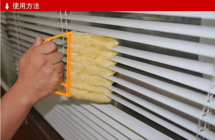 A hand is using an orange tool to clean dust from white horizontal blinds with a yellow dusting brush.