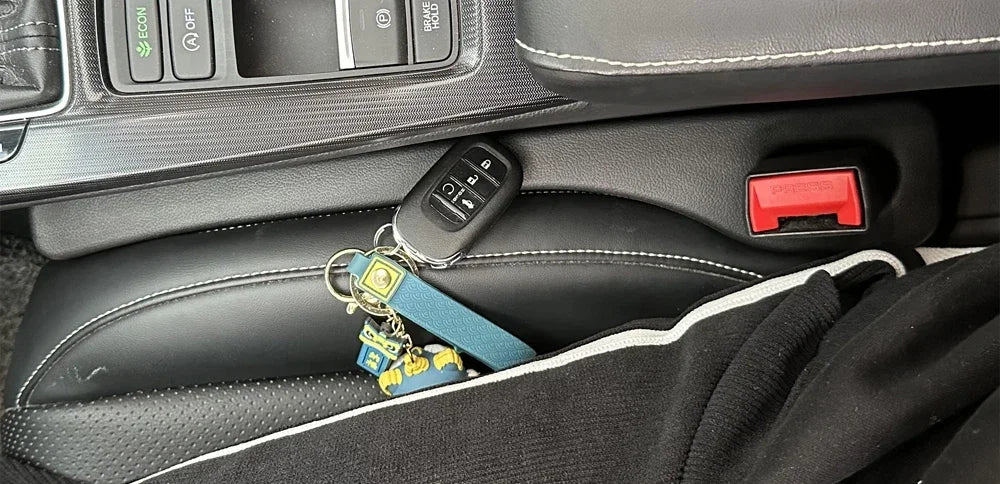 A black universal car seat gap filler and storage organizer with a key fob and various small accessories is shown in a vehicl