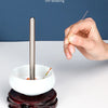 A hand is using a thin stainless steel beading needle to thread beads into a white bowl, demonstrating DIY beading.