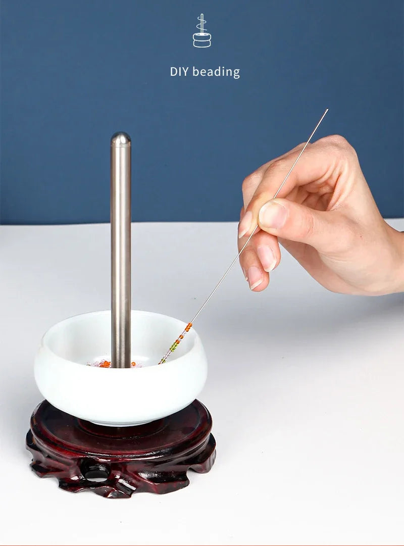 A hand is using a thin stainless steel beading needle to thread beads into a white bowl, demonstrating DIY beading.