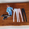 A fashion doll wearing a blue plaid shirt and gray pants is standing on a wooden surface next to various colored stockings.
