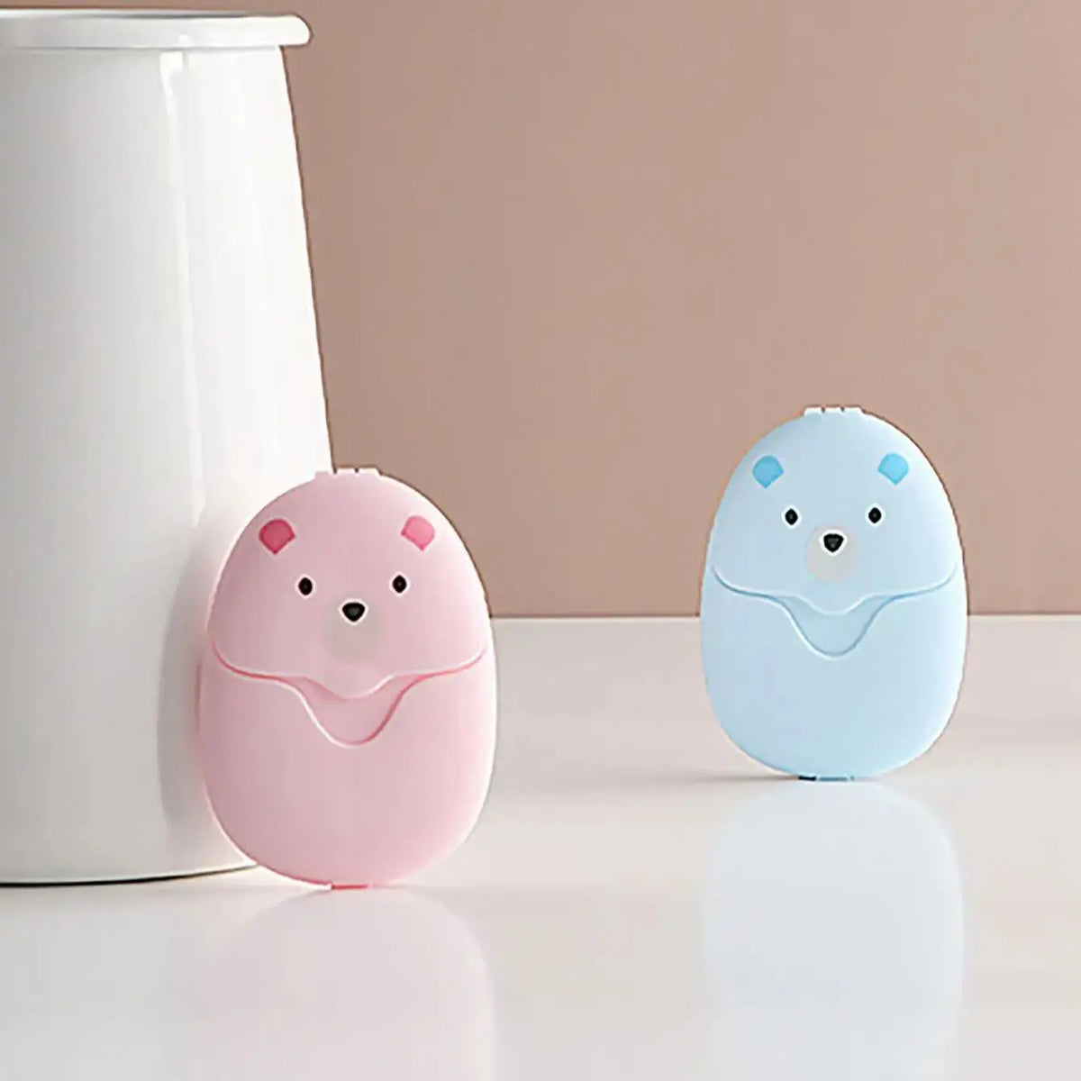 Two round, light pink and blue electronic soap dispensers sit on a white surface next to a white container.