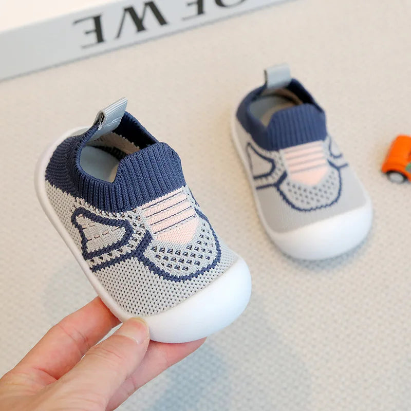 A hand holds a pair of light gray and blue breathable soft-sole baby shoes with non-slip grip.