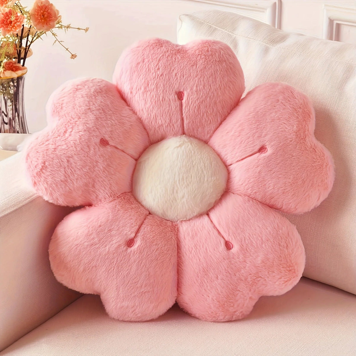 A pink plush daisy flower throw pillow with a white center resting on a light-colored sofa.