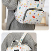 A woman is wearing a white and multi-colored patterned backpack diaper bag with black accents over a gray sweater.