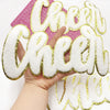 Hand holding a large white chenille letter patch spelling 'Cheer' on a pink background