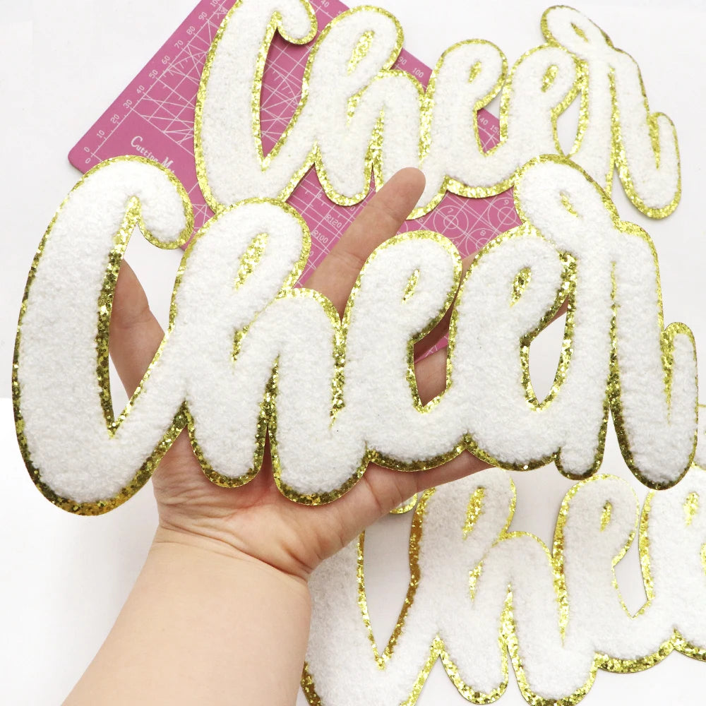 Hand holding a large white chenille letter patch spelling 'Cheer' on a pink background