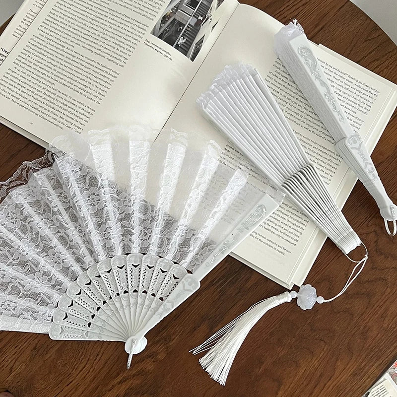 A white vintage lace folding hand fan with a durable handle is displayed next to an open book.