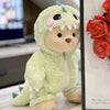 A soft, light green teddy bear doll dressed in a dinosaur jumpsuit outfit.