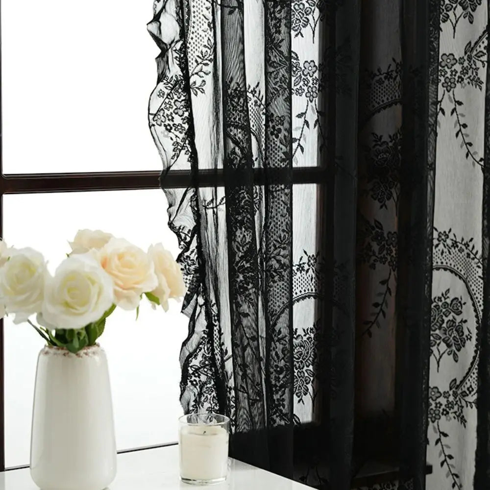 Black and white lace sheer curtains with floral embroidery hanging in a window next to a white vase of roses