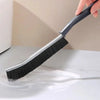 A hand holds a gray grout cleaning brush with black bristles, demonstrating its use on a white surface.