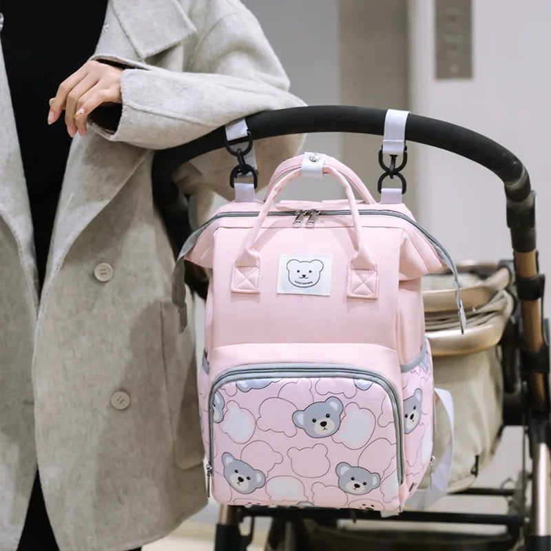 A pink bear print waterproof diaper backpack is shown hanging on a black stroller handle.