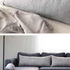 A light gray linen pillowcase is shown folded on a bed, next to a gray sofa.