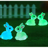 A set of three glowing resin miniature bunny figurines in shades of light green and blue on a dark green surface.