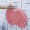 A hand is placing a pink suction cup foot massage mat with exfoliating bristles onto a white tiled wall.