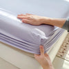 A hand is folding a purple fitted sheet with an elastic band on a white bed.