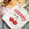 A white canvas tote bag featuring a red cherry print design and text on a wooden surface.