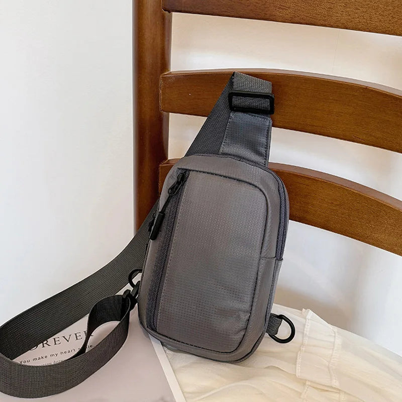 Ein dunkelgrauer, vielseitiger Unisex-Crossbody-Rucksack liegt auf einem hellen Stoff neben einem Holzstuhl.