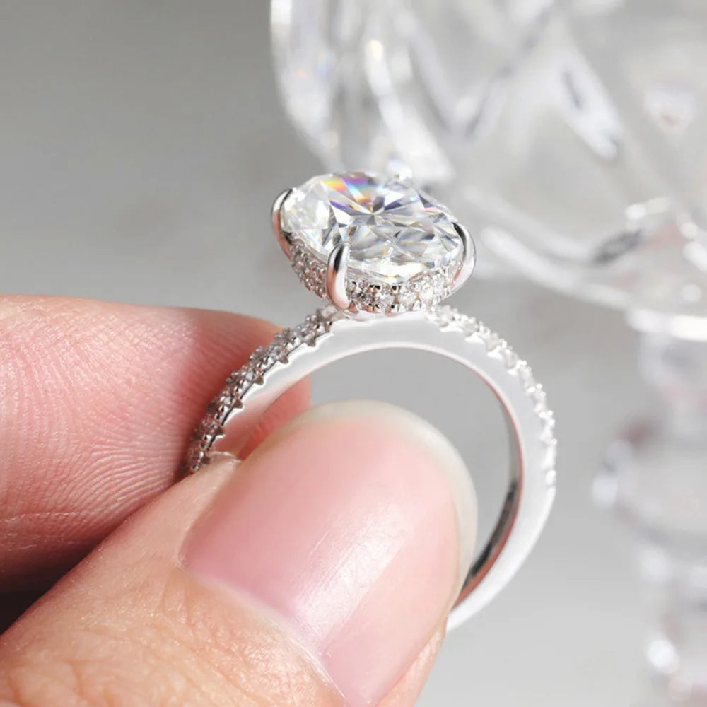 A hand holds a diamond engagement ring featuring an oval moissanite center stone and a hidden halo setting.