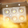 A rectangular LED flood light pod with four illuminated circular light elements is shown against a bright, hazy background.