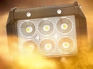 A rectangular LED flood light pod with four illuminated circular light elements is shown against a bright, hazy background.