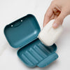 A hand is opening a teal leak-proof travel soap box containing a white bar of soap on a white marble surface.