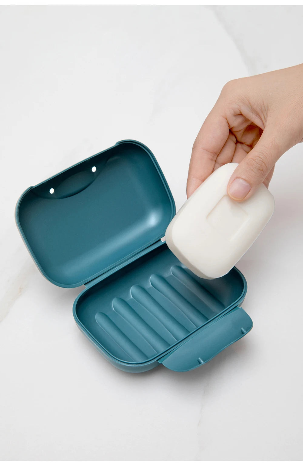 A hand is opening a teal leak-proof travel soap box containing a white bar of soap on a white marble surface.