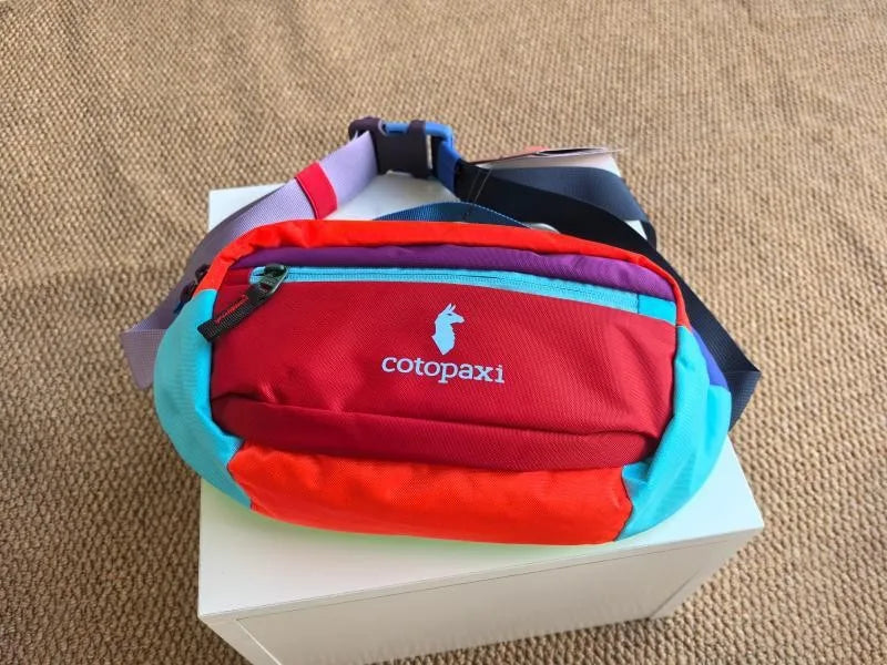 A red and blue crossbody chest bag with a purple strap resting on a white box.