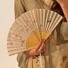 A hand holds an open bamboo and silk folding fan with a floral design against a light background.