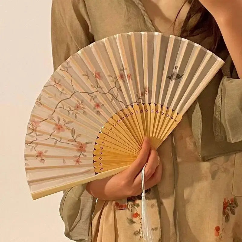 A hand holds an open bamboo and silk folding fan with a floral design against a light background.