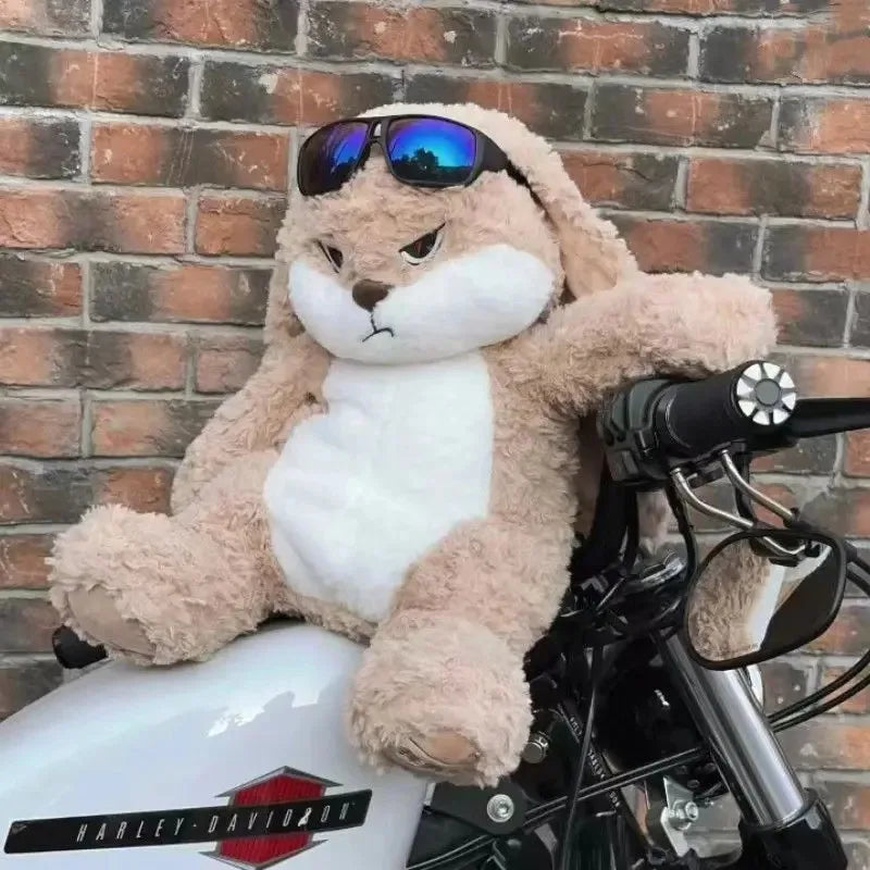 A fluffy brown and white rabbit plush toy wearing sunglasses sitting on a black motorcycle.