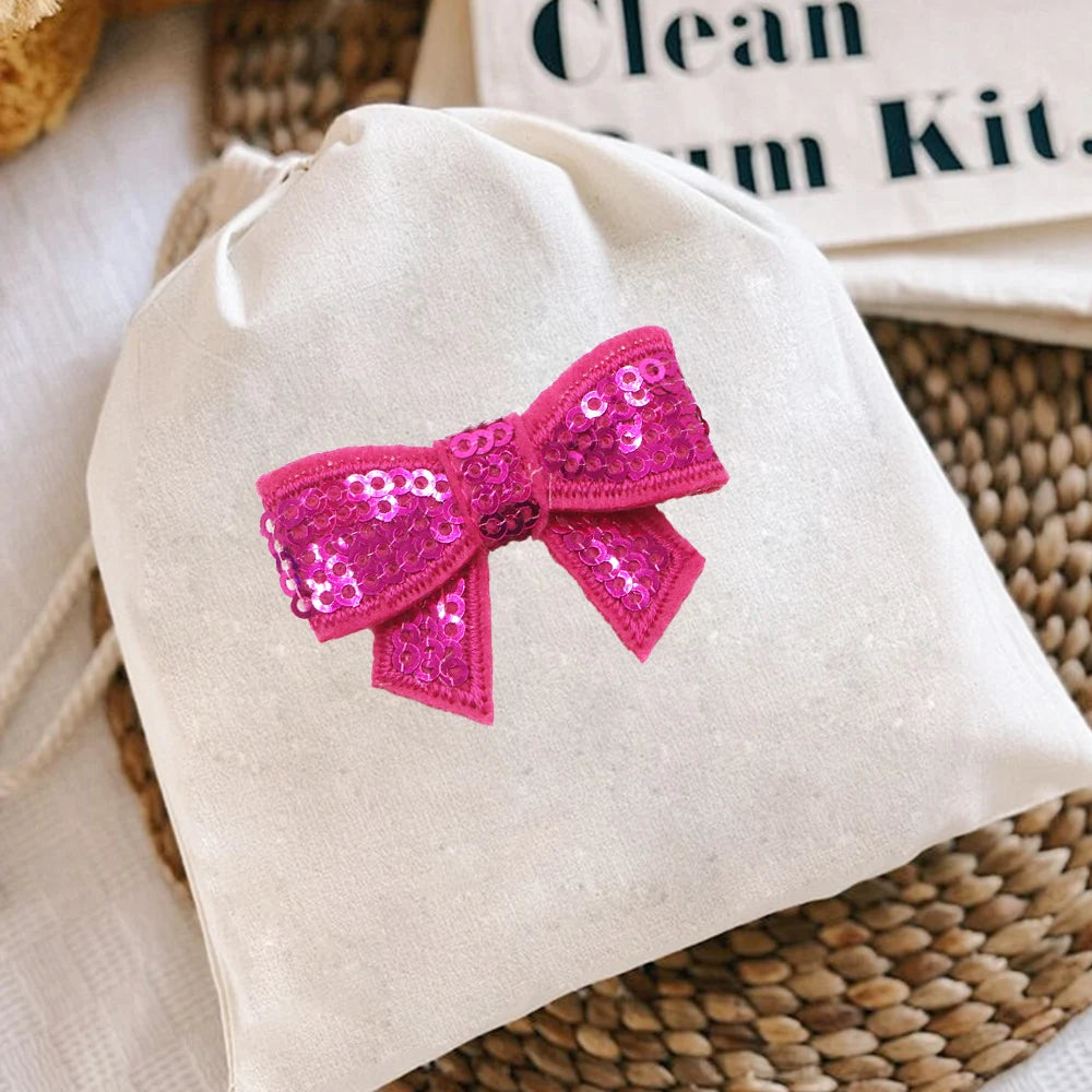 A light beige cloth drawstring bag featuring a bright pink sequin bow patch decoration.