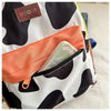 A close-up of a white and black cow print backpack featuring an orange pocket and a brown leather tag.