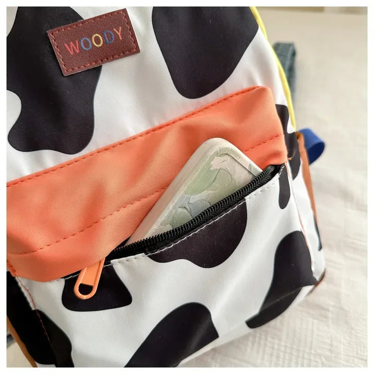 A close-up of a white and black cow print backpack featuring an orange pocket and a brown leather tag.
